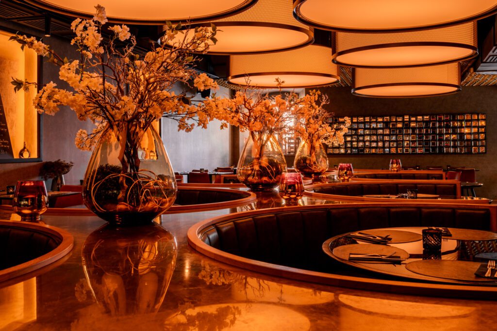 Warm beleuchteter Innenraum mit stilvollen, runden Restaurant-Tischen und großen Blumenvasen