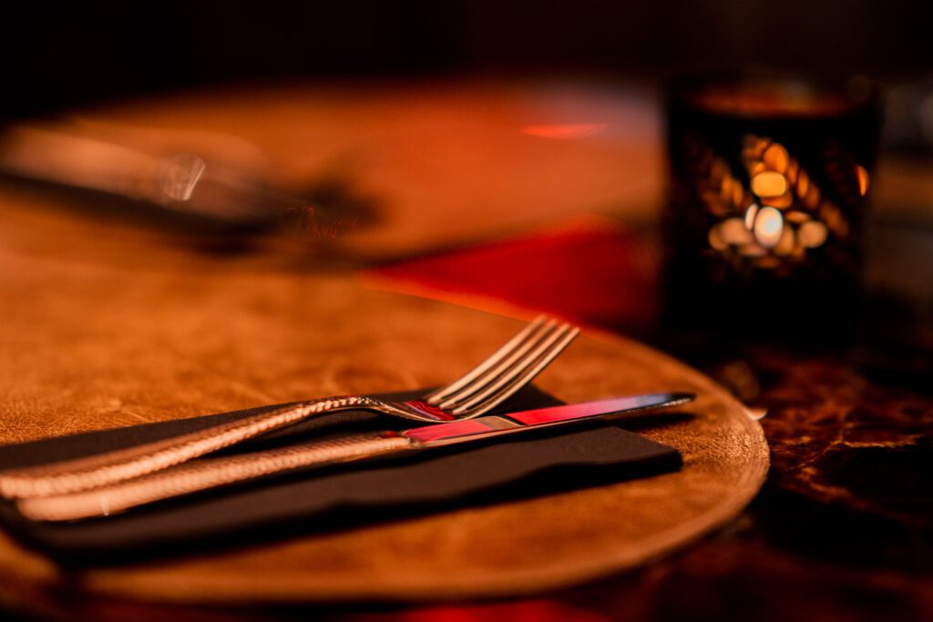 Glänzendes Geschirr liegt auf einem dunklen Tisch mit Leder Untersetzern. Eine Kerze wirft ein warmes Licht auf das Besteck