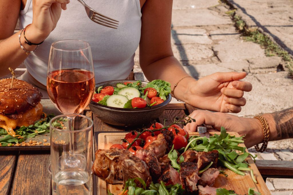 Nahaufnahme eines Tisches mit Essen im Freien, mit Salat, Steak und Burger. Menschen essen im Hintergrund.